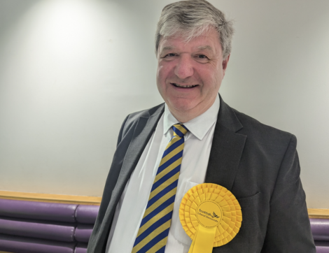A man wearing a suit and tie with a yellow rosette on his chest stands against a white and purple wall, smiling at the camera.