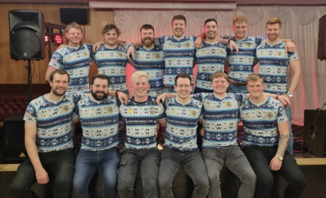 A group of fifteen men wearing matching festive-patterned shirts, posing together inside a room with red and white decorations.