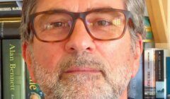A bearded man with glasses is wearing a navy sweater. He stands in front of a bookshelf filled with books.