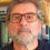 A bearded man with glasses is wearing a navy sweater. He stands in front of a bookshelf filled with books.