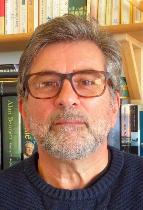 A bearded man with glasses is wearing a navy sweater. He stands in front of a bookshelf filled with books.