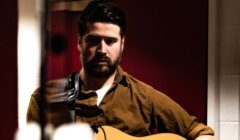 A man with a beard and brown shirt plays an acoustic guitar indoors with a focused expression.