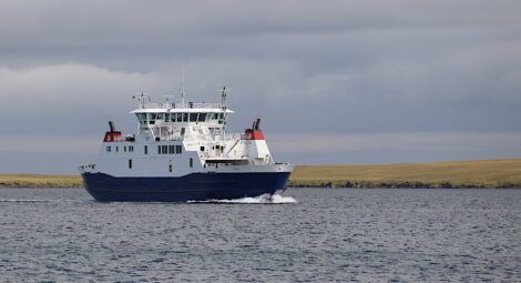 A large blue and white cargo ship sails on a body of water, with grasslands and cloudy skies in the background.