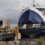 A ferry with its loading ramp lowered docking at a port. The front of the ferry is open, revealing the interior vehicle deck. The port area has yellow railings and a docking structure.