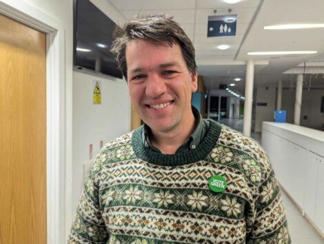 A man wearing a patterned sweater and a "Vote Green" button smiles in a brightly lit indoor hallway.
