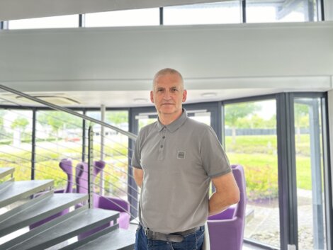 A man in a gray polo shirt stands by a staircase in a modern building with large windows and purple chairs.