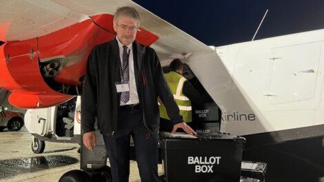 A man stands next to an airplane, holding a black ballot box. An open cargo area under the plane's wing reveals additional ballot boxes being loaded or unloaded.