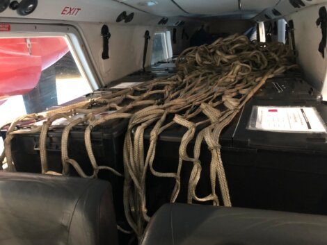 Interior of an aircraft showing a pile of ropes and black cargo containers, with an exit sign visible on the wall.