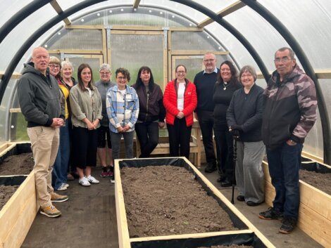 A group of people stands inside a greenhouse structure with raised garden beds filled with soil. They seem to be posing for the photo.