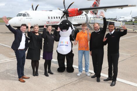 A group of six people, including one in an orca costume, stand in front of a Loganair plane, all raising their hands and smiling.