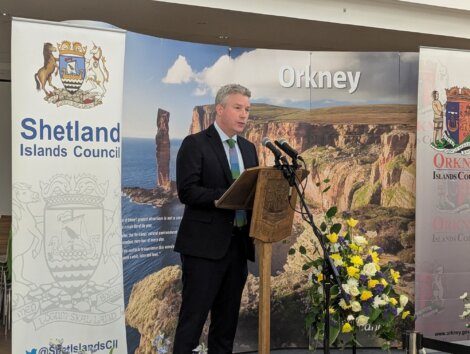 A man stands at a podium with microphones, speaking at an event. Behind him are banners for Shetland Islands Council and Orkney Islands Council, with scenic background images.