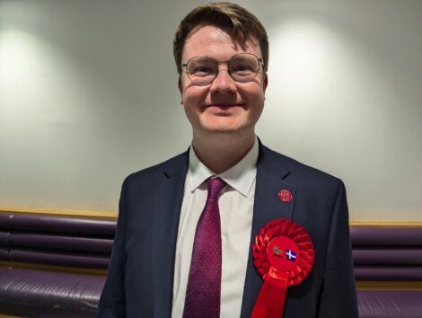 A man wearing glasses and a dark suit with a red ribbon on his jacket is standing in front of a light-colored wall, smiling.