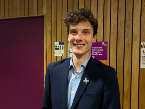 A person wearing a navy blazer and light blue shirt stands in front of a wooden-paneled wall with several posters.