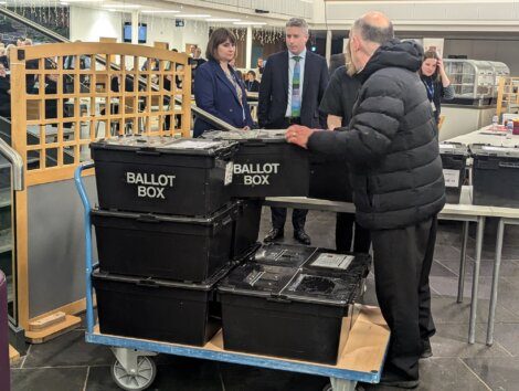A person in a black jacket pushes a cart with stacked ballot boxes labeled "BALLOT BOX." Three individuals observe in the background.