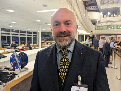 A man with a beard wearing a dark suit, patterned tie, and a name badge stands in a bright, modern indoor setting with people in the background.
