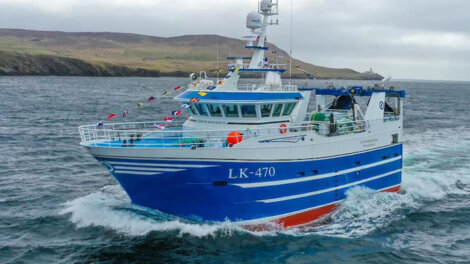 A blue and white fishing boat with the label "LK-470" sails in the ocean near a rocky coastline, decorated with colorful flags.