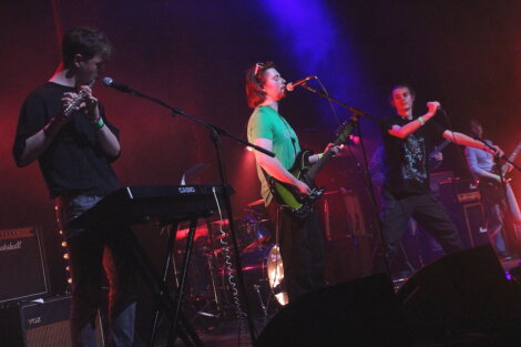 Four musicians perform on stage with various instruments, including a keyboard, guitar, and microphone, under colorful stage lighting.