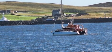A sailboat and a small motorized boat are on the water near a distant shore with green fields and buildings.