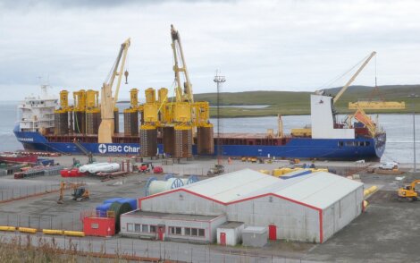 A cargo ship is docked at a port, loaded with large yellow structures and featuring cranes. Buildings and equipment are seen in the foreground, with a body of water and land in the background.
