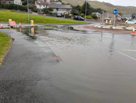 Flooded roundabout with traffic cones and a road closed sign indicating the area is impassable due to heavy rain. Surrounding residential houses and lawns are visible in the background.