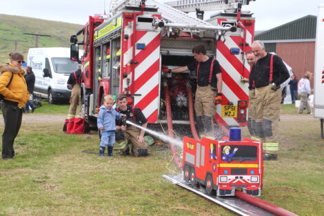 Firefighters demonstrate using a fire hose with a small child, aiming water at a mini fire truck model, with a full-sized fire truck in the background. Several people observe the scene.