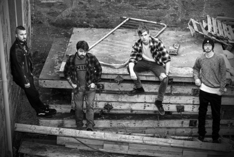 Black and white photo of four men outdoors, casually standing and sitting on stacks of wooden pallets and construction debris, with an old metal bed frame in the background.