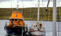 A lifeboat assists a sailboat on calm waters with crew members working on both boats; landmass and clouded sky in the background.
