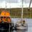 A lifeboat assists a sailboat on calm waters with crew members working on both boats; landmass and clouded sky in the background.