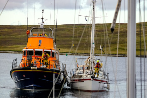 A lifeboat assists a sailboat on calm waters with crew members working on both boats; landmass and clouded sky in the background.