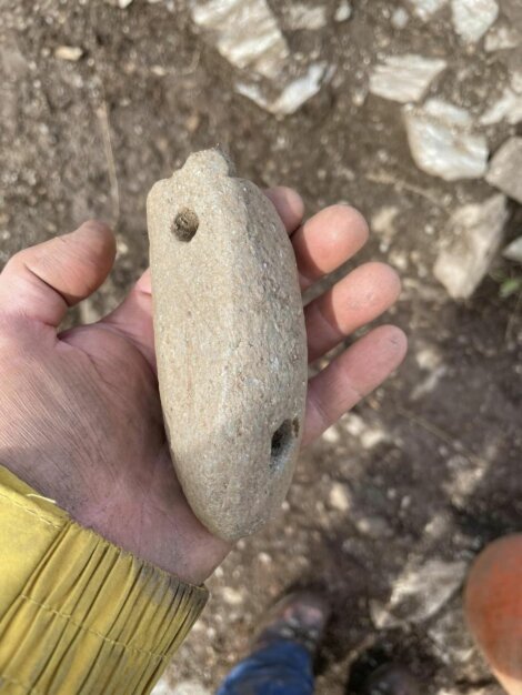 A person wearing a yellow sleeve holds a rough, elongated stone object with two holes drilled through it, against a backdrop of soil and rocks.