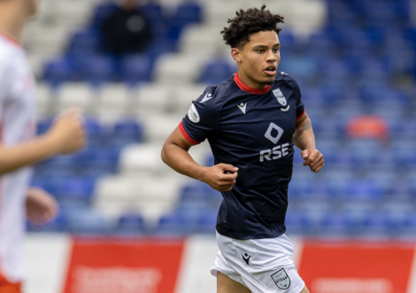A soccer player wearing a dark blue jersey with white shorts is running on a field during a match. Another player is partially visible on the left side of the image.