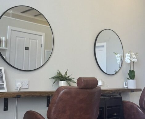 Two brown leather chairs facing two round mirrors in a minimalist salon setting, with potted plants and a monochrome photograph on a wooden shelf.
