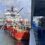 A red and white supply vessel is docked at a port next to a concrete pier, with various equipment and personnel visible on board. The sky is partly cloudy.
