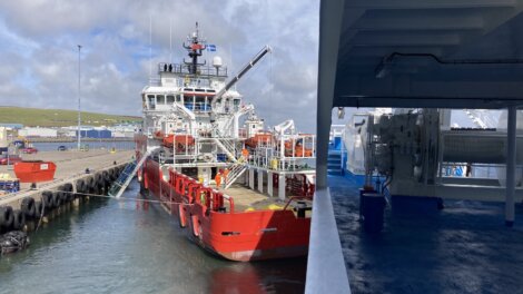 A red and white supply vessel is docked at a port next to a concrete pier, with various equipment and personnel visible on board. The sky is partly cloudy.