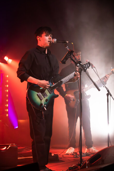A guitarist and a bassist perform on stage under reddish lighting, both dressed in dark clothing. The guitarist sings into a microphone while playing. Equipment and stage lights are visible in the background.