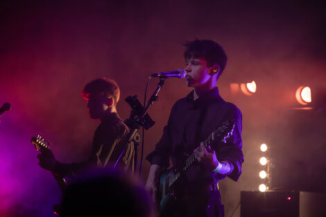 Two musicians perform on stage with guitars, one singing into a microphone. The background is lit with colorful stage lights and smoke effects.