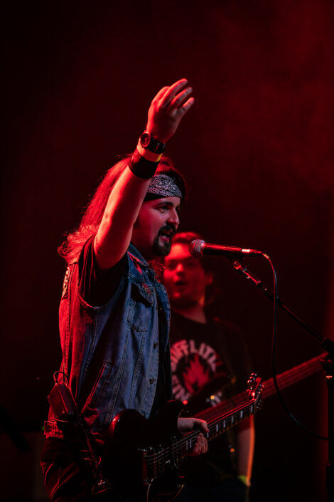 A musician with long hair, wearing a bandana and denim vest, plays an electric guitar while singing into a microphone on a dimly lit stage with red lighting. Another musician is in the background.