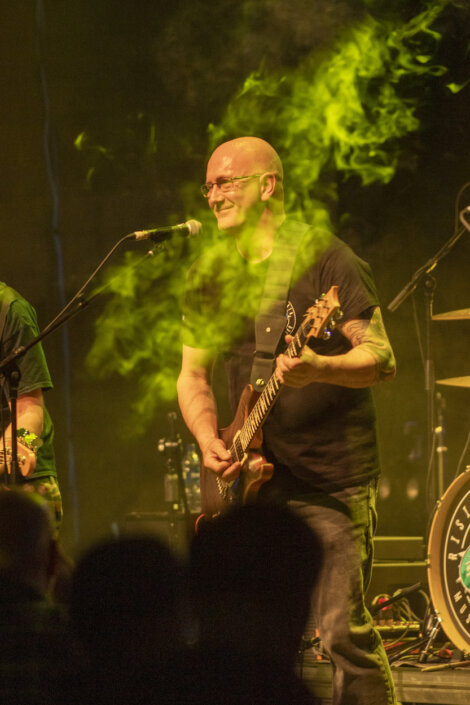 A musician plays an electric guitar and sings into a microphone on a stage with green lights and smoke effects, while a drum set is visible in the background.