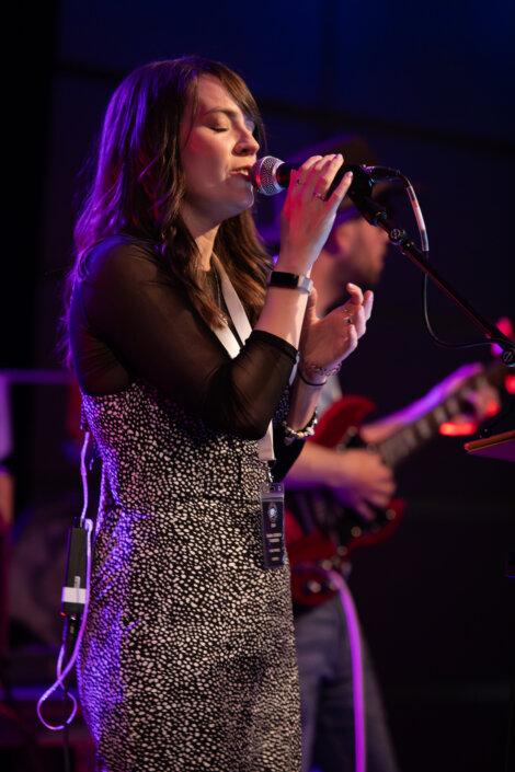 A woman in a black and white patterned dress sings into a microphone on stage with a guitar player in the background.