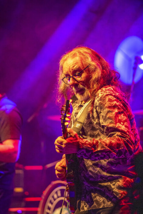 A man with long hair and glasses plays an electric guitar on stage, illuminated by colorful lights.