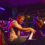 A band performs on stage with a guitarist singing into a microphone, a pianist playing a keyboard, a bassist, and a drummer. The scene is lit with purple stage lights.