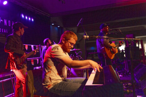 A band performs on stage with a guitarist singing into a microphone, a pianist playing a keyboard, a bassist, and a drummer. The scene is lit with purple stage lights.