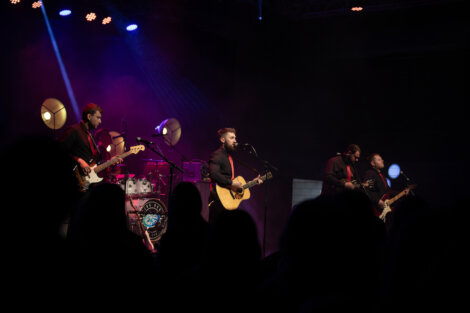 A band performs on stage with instruments, including guitars and drums, under colorful stage lighting. Audience members are visible in silhouette in the foreground.