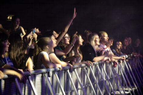 A crowd of people stand behind a barrier at a concert. Some are raising their hands and cheering, while others are looking towards the stage. The area is dimly lit with concert lighting.