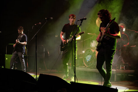 A four-member band performs on stage under green and yellow lighting. Three members play guitars while the fourth is on a drum set.
