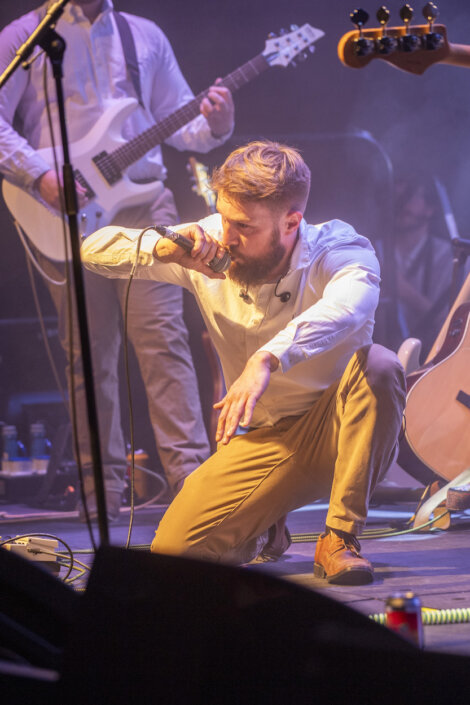 A bearded man kneels on stage, holding a microphone close to his mouth. Behind him, other musicians play guitars under stage lighting.