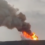 A large plume of smoke and flames is seen rising from a hill, indicating a significant fire or explosion with cloudy skies in the background.
