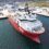 A large red and white research vessel is docked at an industrial harbor, with several buildings, shipping containers, and equipment in the background.
