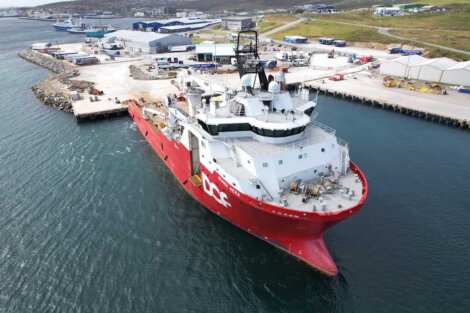 A large red and white research vessel is docked at an industrial harbor, with several buildings, shipping containers, and equipment in the background.
