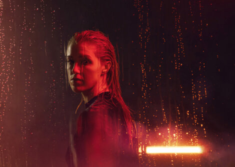 A person with wet hair stands against a dark background with red and orange lights and water droplets.